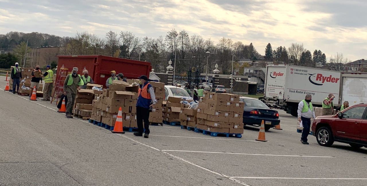 Volunteers in safety vests beside pallets of boxes, with cars lined up along a drive-through pickup lane.